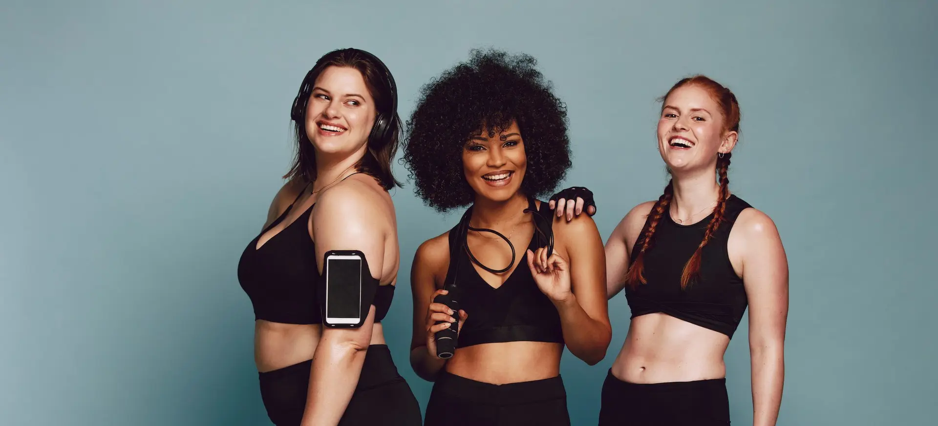Tre donne in abbigliamento sportivo ridono insieme davanti a uno sfondo grigio-azzurro; una indossa le cuffie e lo smartphone al braccio